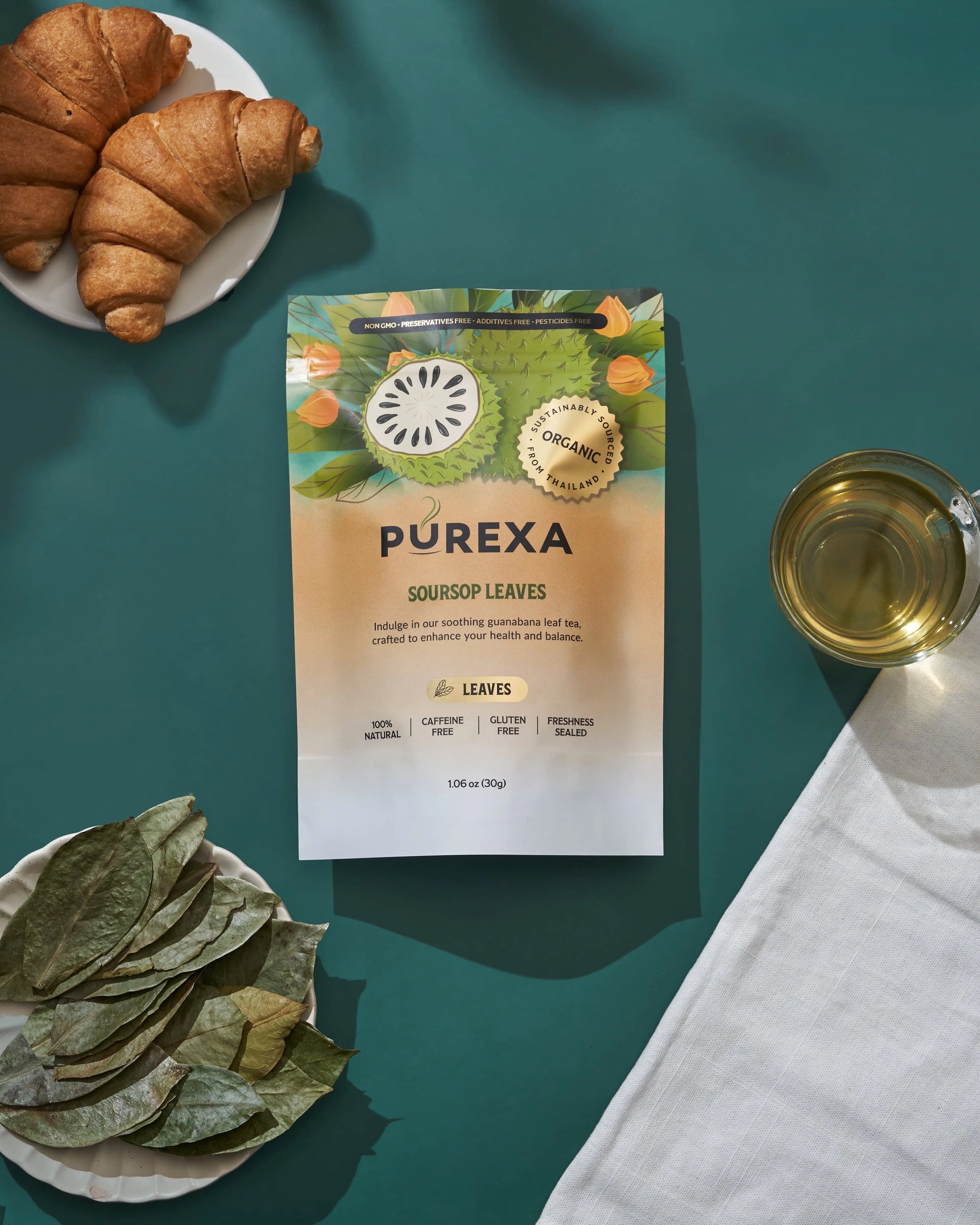 Overhead view of PUREXA soursop leaves pouch with dried leaves, brewed tea, and croissants